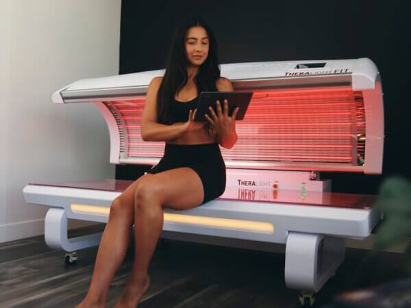 Woman sitting in a red light therapy bed for gyms looking at ipad