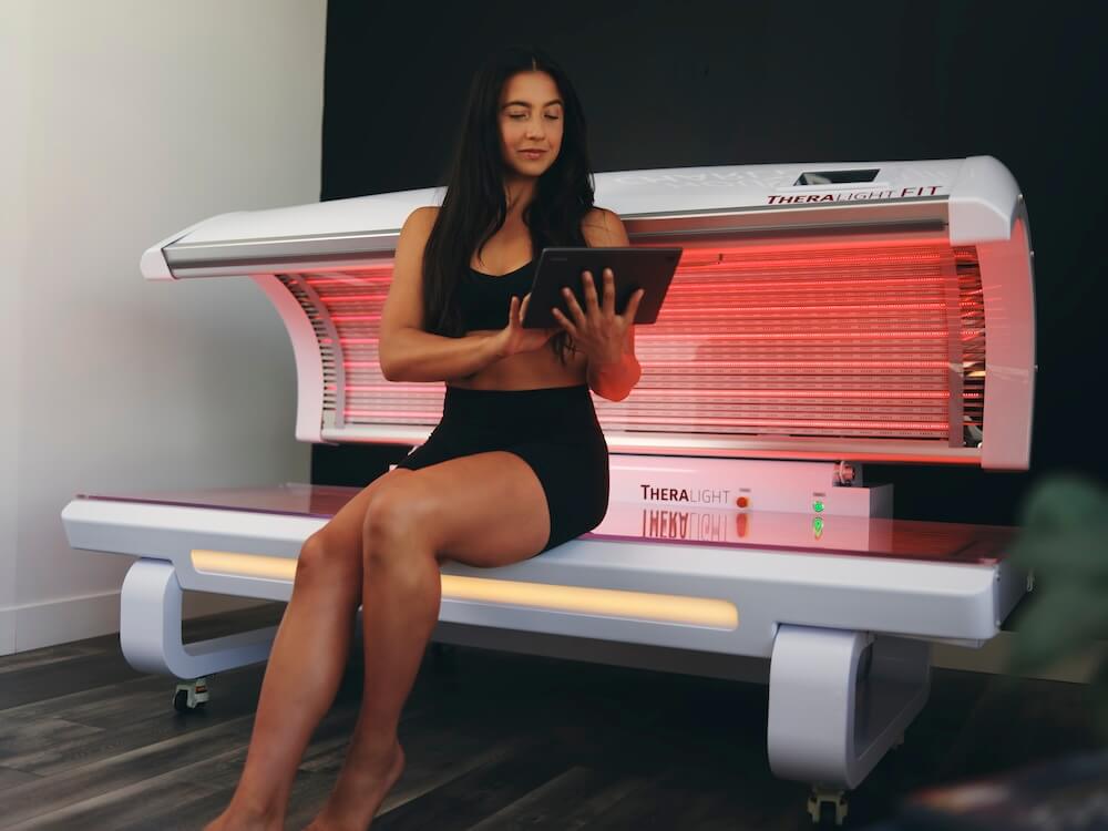 Woman sitting in a red light therapy bed for gyms looking at ipad