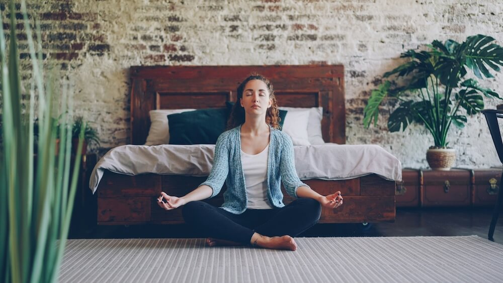 Woman meditating in front of her bed showing that TheraLight Enhances Your Wellness Routine
